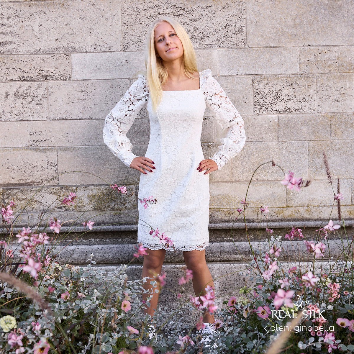 pige står bag nogle lyserøde blomster i en hvid konfirmationskjole med lange luftige ærmer og manchet. Kjolen er en bodycon kjole og den er syet i stretchblonde med blomstermotiv