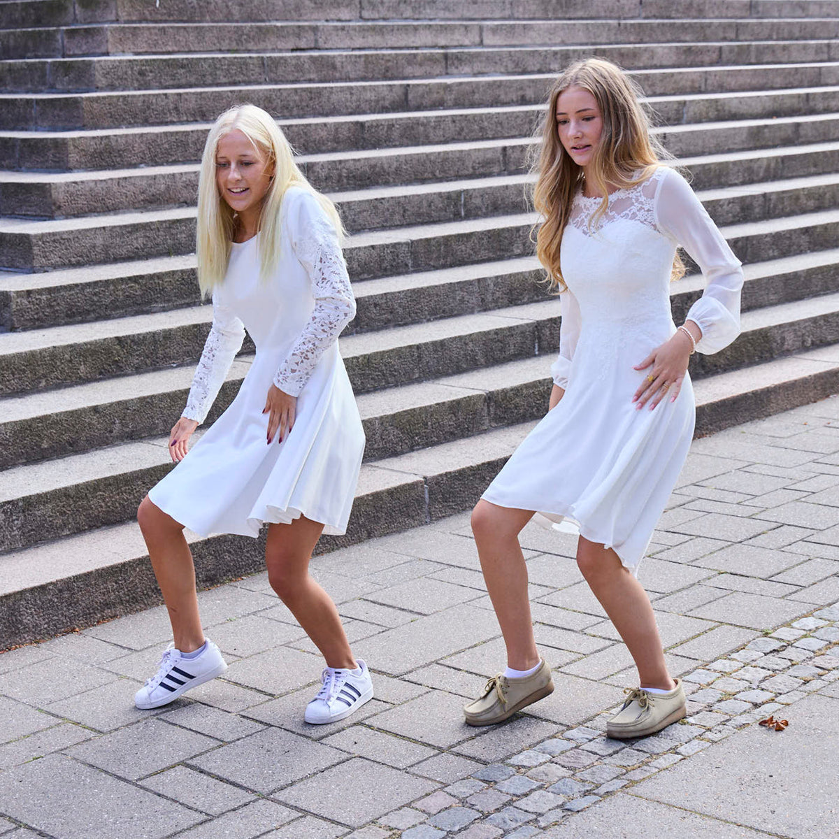 To piger i hvide konfirmationskjoler med lange ærmer og blonder, danser foran en trappe